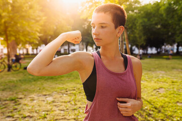 teenage girl in sports shorts and top demonstrates muscles on arm in public park in sun. Healthy active lifestyle, sports, outdoor fitness. Useful training, relaxation, muscle stretching.