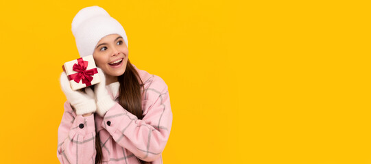 curious kid in hat and mittens hold gift box on yellow background, new year. Banner of christmas child girl, studio kid winter portrait with copy space.