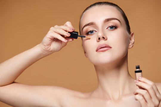 woman applying concealer under her eyes on beige background
