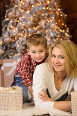 little blond boy hugging his mom lying on the floor, near gift boxes, against the backdrop of a christmas tree