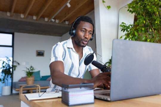 Happy african american man sitting at table in kitchen, using laptop and making vlog