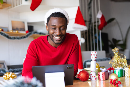 Happy african american man wearing santa hat, using tablet for video call - Powered by Adobe