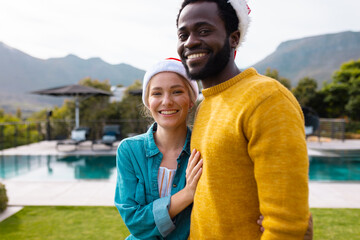 Portrait of happy diverse couple in santa hats to celebrate christmas smiling outdoors