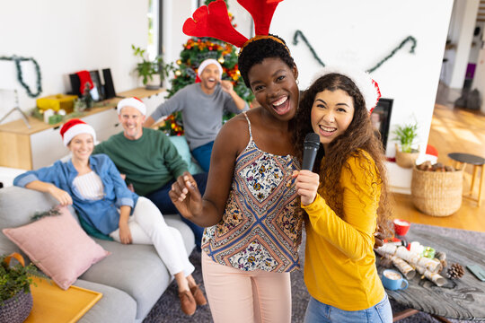 Image of happy two diverse female friends singing karaoke celebrating christmas at home with friends