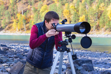 Adult attractive man in sportswear looking through telescope on bank of mountain river background.