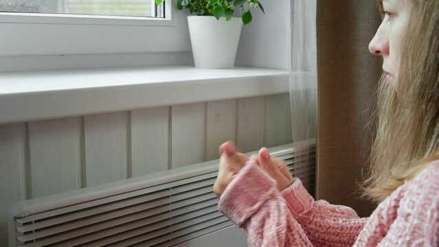 Young frozen woman wearing a sweater shaking and freezing for winter cold with shock expression on face. Girl is warming up hands with breath over electric heater. Discomfort spending time at home