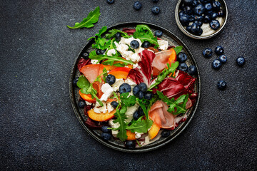 Fresh salad with arugula, radicchio, sweet peaches, smoked pork ham, feta cheese and blueberries. Black stone table background, top view, close up