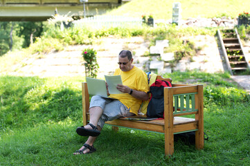 Fototapeta premium Middle-aged man man sitting on the wooden bench in park and using laptop, working with documents outdoors