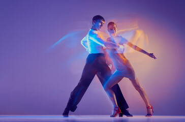 Young graceful dancers, flexible man and woman dancing ballroom dance isolated on gradient blue purple background in neon mixed light.