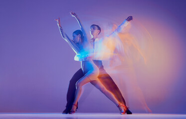 Beautiful woman and slim man, couple of ballroom dancers in stage attires dancing isolated on purple background in neon mixed light.