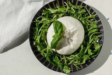 Traditional Italian, fresh, soft burrato ball of young mozzarella cheese with cream on a round dark plate with salad greens on a light table. Flat lay close-up