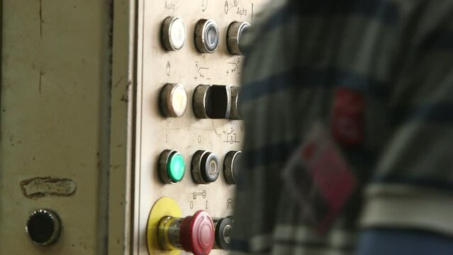 A pair of gloved hands of a manual worker switching on an orange button, turning and adjusting the knobs on the machine control station. 