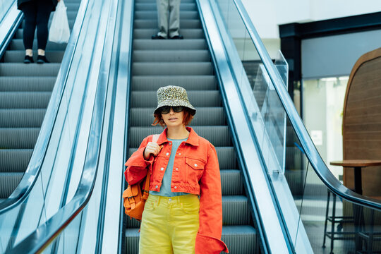 Redhead hipster young woman in bright clothes, sun glasses and bucket hat with backpack bag standing on escalator going down by electric stair elevator in the shoping center. Selective focus.