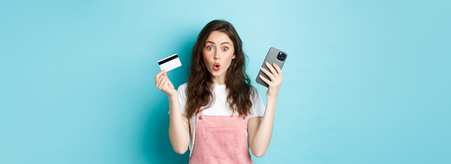 Surprised young woman say wow, stare excited at camera, holding mobile phone and plastic credit...