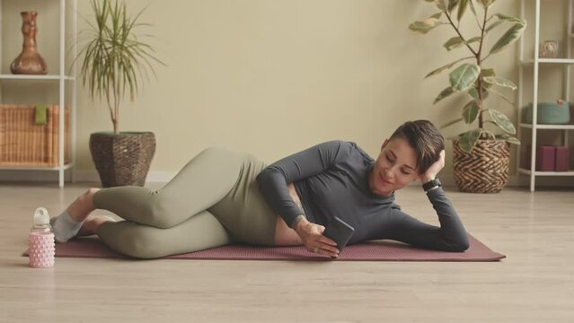Contemporary pregnant woman texting on smartphone while lying relaxedly on yoga mat at home after practice