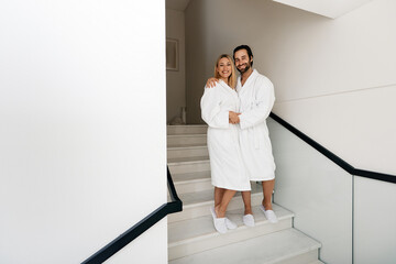 Spa family day. Handsome man with his beautiful wife in white bathrobes enjoying spa weekend while honeymoon walking at wellness center
