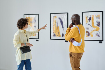 Young woman discussing paintings on wall together with African man while they visiting art gallery - obrazy, fototapety, plakaty