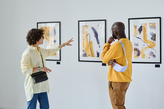 Young woman discussing paintings on wall together with African man while they visiting art gallery