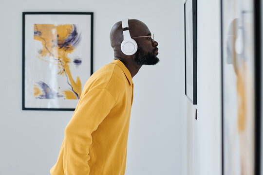 African young man in wireless headphones examining painting on the wall at gallery