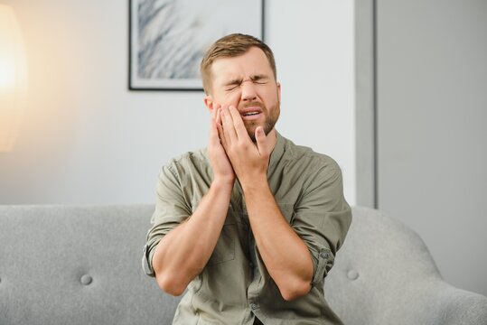 Man suffering from strong toothache. Upset male patient with inflamed nerve, tooth abscess, or teeth sensitivity sitting on sofa and touching cheek with grimace of pain.