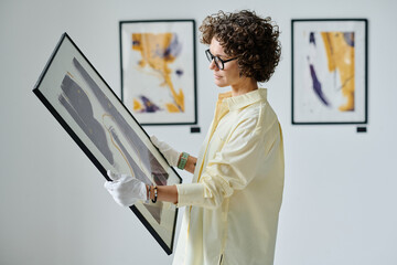 Young woman working with art at gallery, she standing and examining picture in frame in her hands - obrazy, fototapety, plakaty