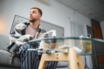 Obraz premium It's cold at home in wintertime. Man freezing in his house in winter because of broken thermostat. young guy wrapped in woolen plaid shivering while sitting on sofa in living room interior