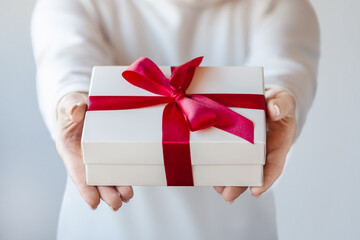 A woman holds in her hands a white box tied with a red ribbon with a bow. Close-up of female hands holding a gift box.