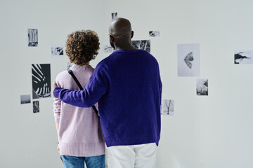 Rear view of multiethnic couple visiting photo gallery, they embracing and watching at photos on...