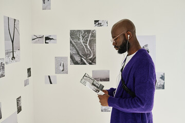 African man in headphones reading booklet while examining pictures at art gallery