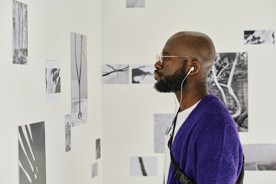 Side view of African man in headphones enjoying modern art at gallery looking at pictures on wall