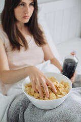 Woman in bed, drinking cola and eating chips while watching TV. Junk food concept.
