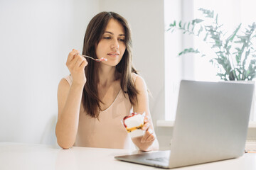 The concept of working at home. Young woman with a laptop in the kitchen, working with pleasure and eating delicious ice cream.