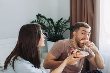 Happy young man and woman relaxing on bed at home watching TV together and eating donuts and fruit. The concept of rest and junk food.