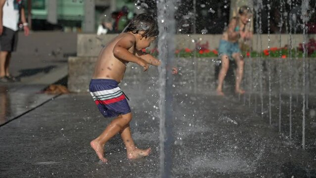 Little Boy Playing At City Water Jets In Super Slow Motion. Child Plays Outdoors During Hot Summer Daylight