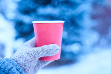 Cup in hand in winter with a hot drink, mock up for your text, winter walk with hot tea in gloved hand, red paper cup eco on a blue background. High quality photo