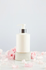 Obraz na płótnie Canvas Dispenser white bottle on marble podium with tender pink roses and rose petals. Natural beauty product based on rose flowers, fermented cosmetic. Soft focus style, copy space, vertical format
