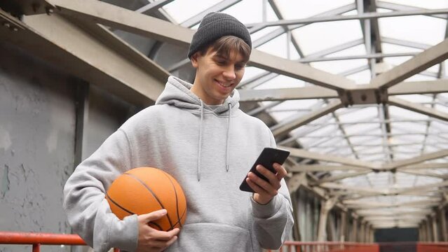 Young man basketball player with headphones holding ball using smartphone after training. Urban background.