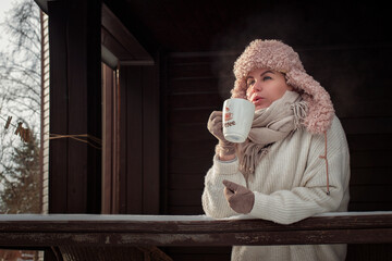A girl in a pink hat drinks tea on the veranda of a wooden house in winter - Powered by Adobe