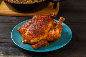Whole baked chicken carcass with a crust on a platter on a wooden table next to a frying pan with noodles.