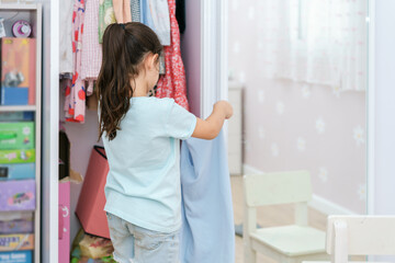 Beautiful little girl with long black hair brings wardrobe order, puts everything in its place in closet. Wardrobe with child's clothing.