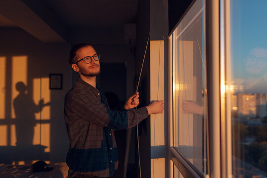 Portrait of young smiling man closes window rolled jalousies in sunny day at sunset. Wearing eyeglasses.