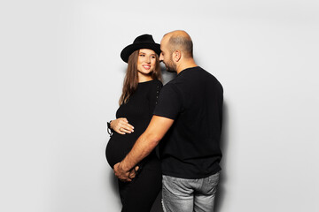 Studio portrait of young parents, pregnant woman wearing black hat and dress, man touching the belly on white background.