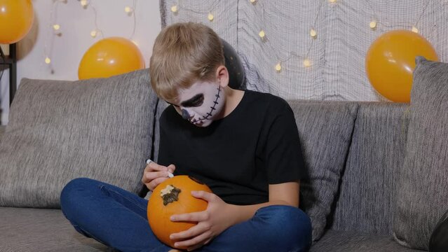 A boy in skeleton makeup paints a pumpkin for Halloween while sitting on the couch at home. Child is Ready for the trick or treat holiday.