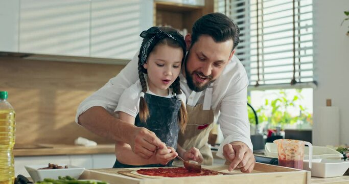 Busy handsome father in apron spending time with adorable little daughter in kitchen dressing homemade pizza in modern kitchen.Curious kid having fun enjoying time with young dad at weekeends.