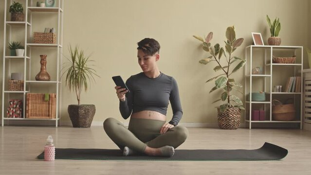Young pregnant woman with short haircut and tattoo on belly texting on smartphone and smiling while having yoga meditation at home