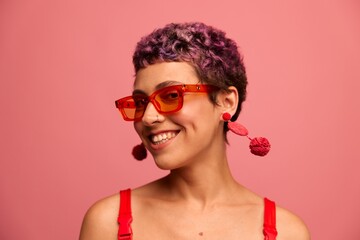 Fashion portrait of a woman with a short haircut in colored sunglasses with unusual accessories with earrings smiling on a pink bright background