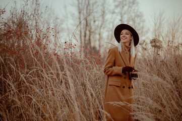 Happy smiling fashionable blonde woman wearing trendy autumn outfit with classic brown coat, hat, suede gloves, beige turtleneck, posing outdoor. Copy, empty space for text