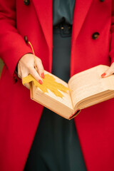 A European girl in a red coat holds an open book with a yellow maple leaf in her hands