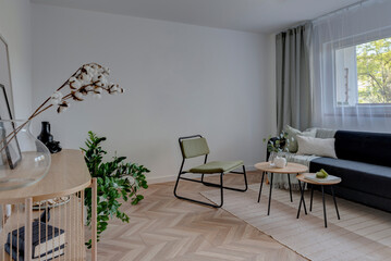 Interior of living room with wooden parquet, window with curtains, white wall and comfortable sofa and armchair with table. Cozy room in apartment.