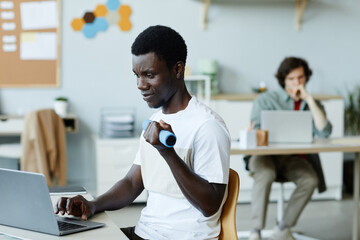 Portrait of young black man holding dumbbell in hand while using computer at workplace, office...